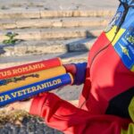 Close-up of three colorful batons held by a person in a bright red, blue, and yellow jacket; inscriptions read 'INVICTUS ROMANIA' and 'ȘTAFETA VETERANILOR' with stone steps in the background.