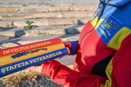 Close-up of three colorful batons held by a person in a bright red, blue, and yellow jacket; inscriptions read 'INVICTUS ROMANIA' and 'ȘTAFETA VETERANILOR' with stone steps in the background.