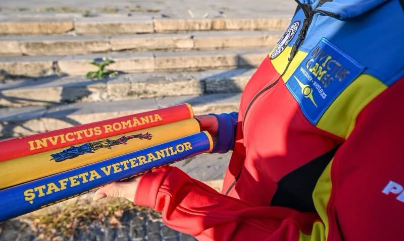 Close-up of three colorful batons held by a person in a bright red, blue, and yellow jacket; inscriptions read 'INVICTUS ROMANIA' and 'ȘTAFETA VETERANILOR' with stone steps in the background.