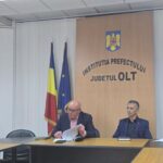 Two men seated at a conference table in a government office, with Romanian and EU flags beside a wooden panel reading 'Institutia Prefectului Județul Olt' and a crest above it.