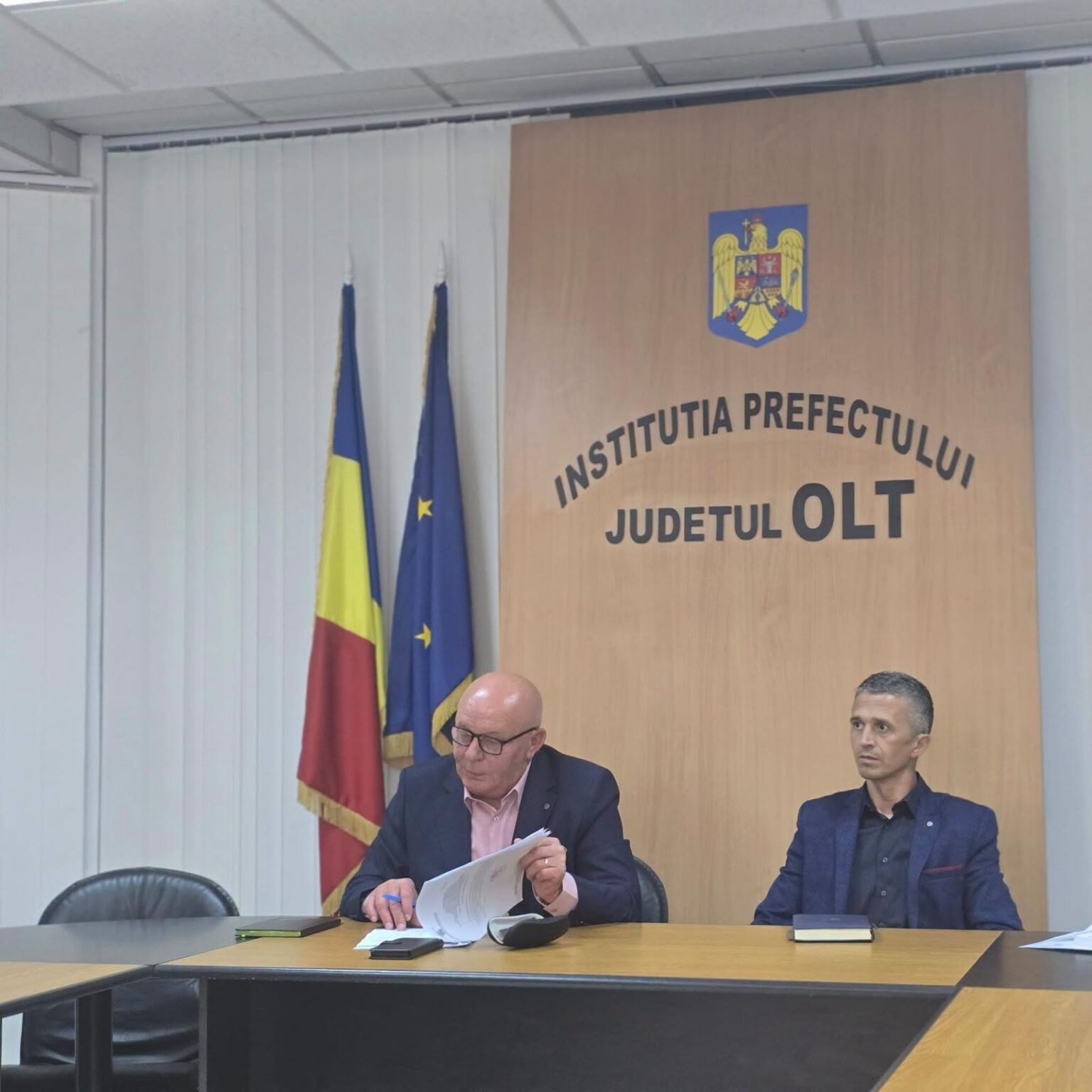 Two men seated at a conference table in a government office, with Romanian and EU flags beside a wooden panel reading 'Institutia Prefectului Județul Olt' and a crest above it.