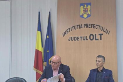 Two men seated at a conference table in a government office, with Romanian and EU flags beside a wooden panel reading 'Institutia Prefectului Județul Olt' and a crest above it.