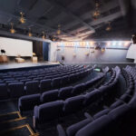 Empty auditorium with a stage, white screen, and rows of blue seats; starburst light fixtures hang from the ceiling above the stage.