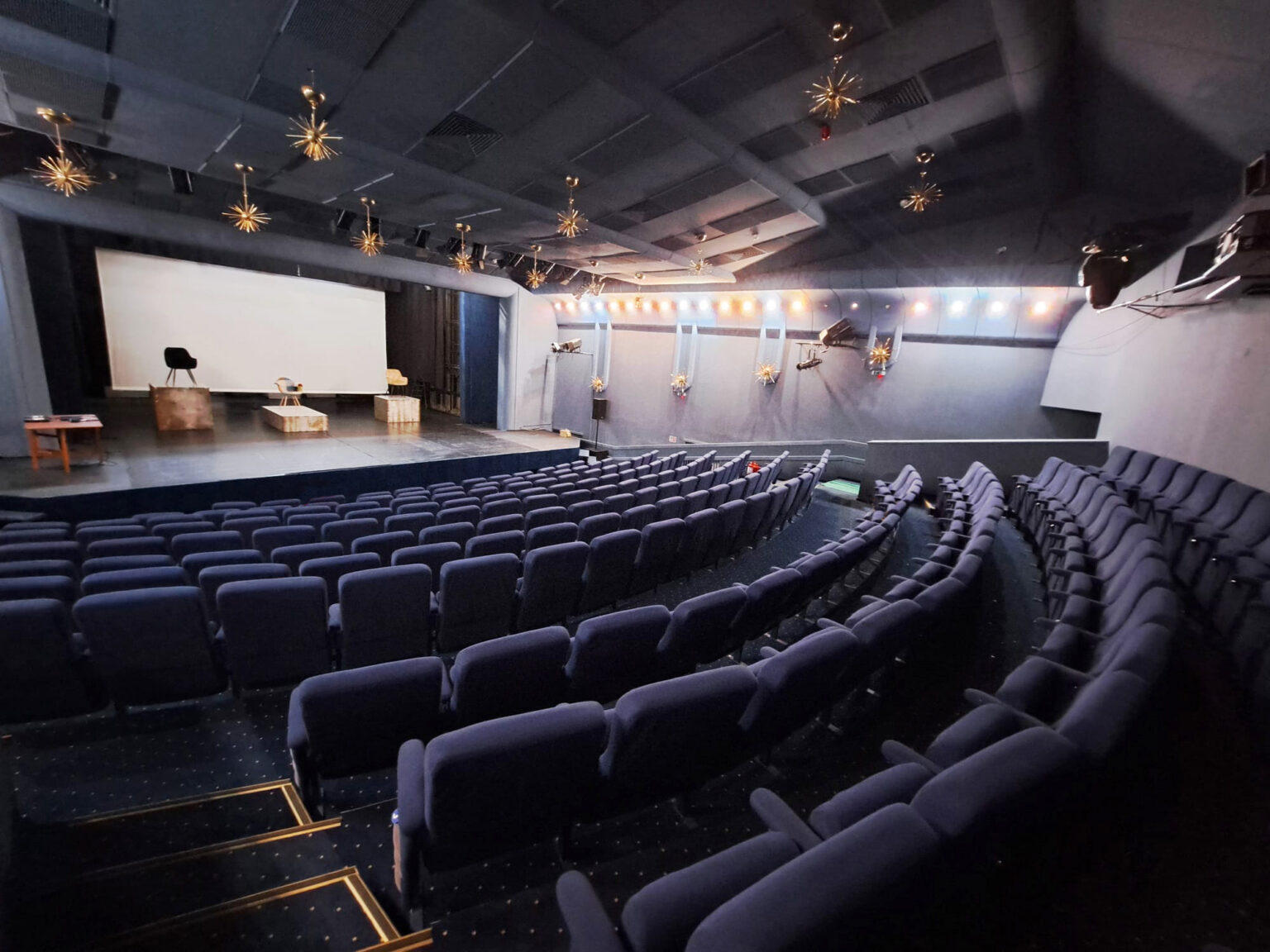 Empty auditorium with a stage, white screen, and rows of blue seats; starburst light fixtures hang from the ceiling above the stage.