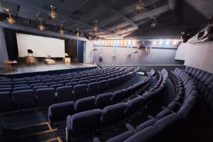 Empty auditorium with a stage, white screen, and rows of blue seats; starburst light fixtures hang from the ceiling above the stage.