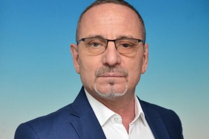 Head-and-shoulders portrait of a middle-aged man with glasses in a navy suit against a blue gradient background, looking at the camera.