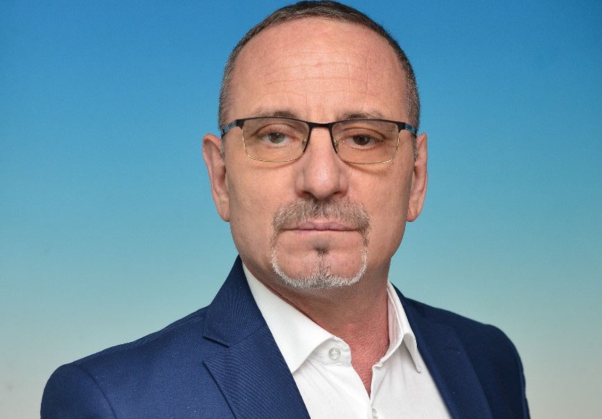 Head-and-shoulders portrait of a middle-aged man with glasses in a navy suit against a blue gradient background, looking at the camera.