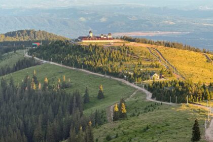 Church complex with red roofs and domed towers atop a hill, surrounded by pine forest and winding roads at sunset.