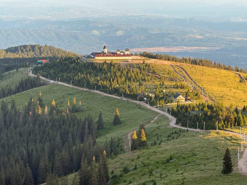 Church complex with red roofs and domed towers atop a hill, surrounded by pine forest and winding roads at sunset.