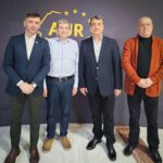 Group portrait of four men standing side by side indoors, dressed in blazers and smart casual wear, in front of a dark backdrop with a yellow ADR logo and stars.