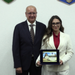 Two professionals in formal attire stand side by side; the woman holds a framed award plaque.