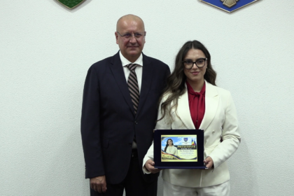 Two professionals in formal attire stand side by side; the woman holds a framed award plaque.