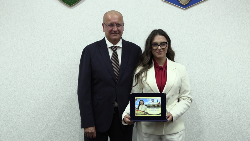 Two professionals in formal attire stand side by side; the woman holds a framed award plaque.
