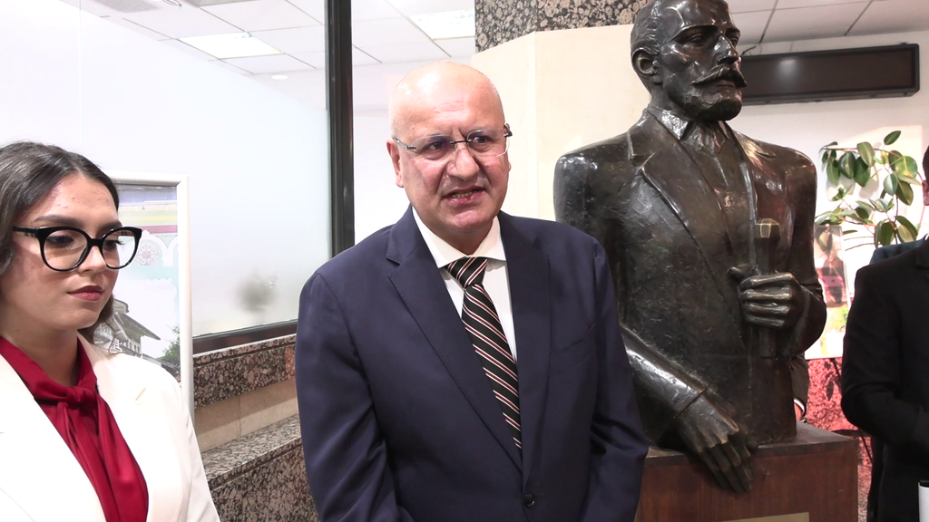 Three people in a lobby: a woman with glasses on the left, a bald man in a navy suit speaking, and a bronze statue of a man in a suit on the right.