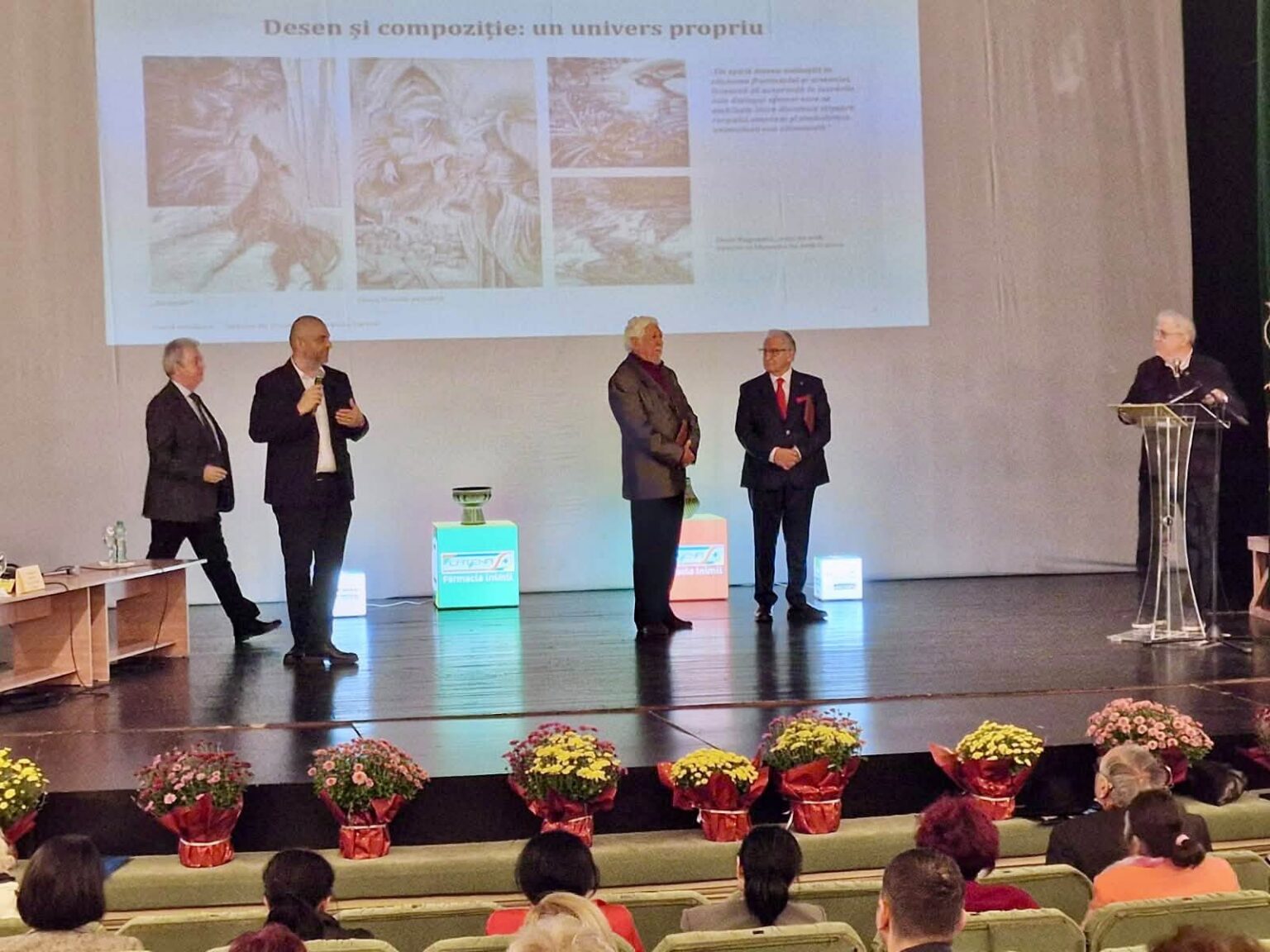 Five men in suits on a stage giving a presentation, with a projected slide behind them and flowers at the front edge of the stage.
