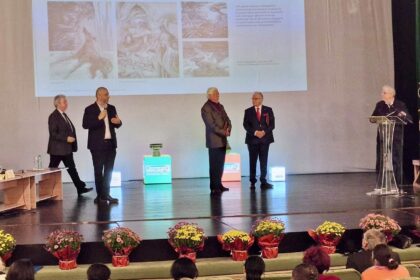 Five men in suits on a stage giving a presentation, with a projected slide behind them and flowers at the front edge of the stage.